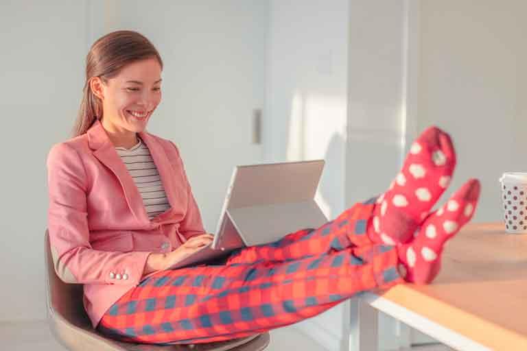 A woman dressed in blouse and jacket for work, but pajama pants on her legs is smiling and looking at the laptop on her lap. Her feet are propped up on the table.
