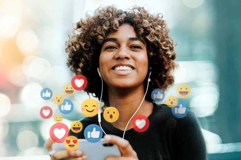 A young woman with earbuds in and emojis floating in the air around her device